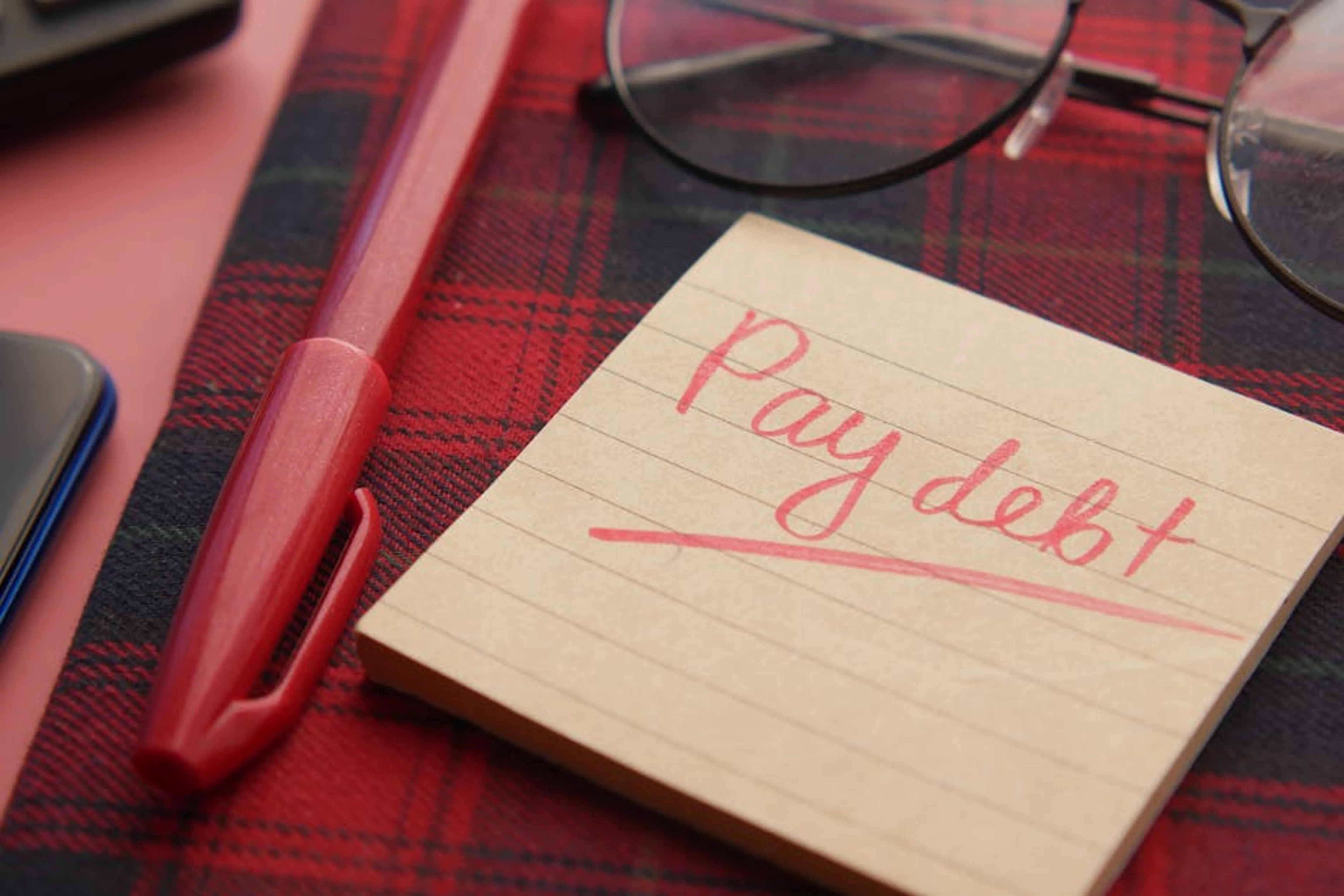 Close-up of a note reading 'Pay debt' next to a red pen on a plaid fabric, emphasizing financial reminders. A person looking stressed while reviewing financial documents and bills, symbolizing the impact of debt