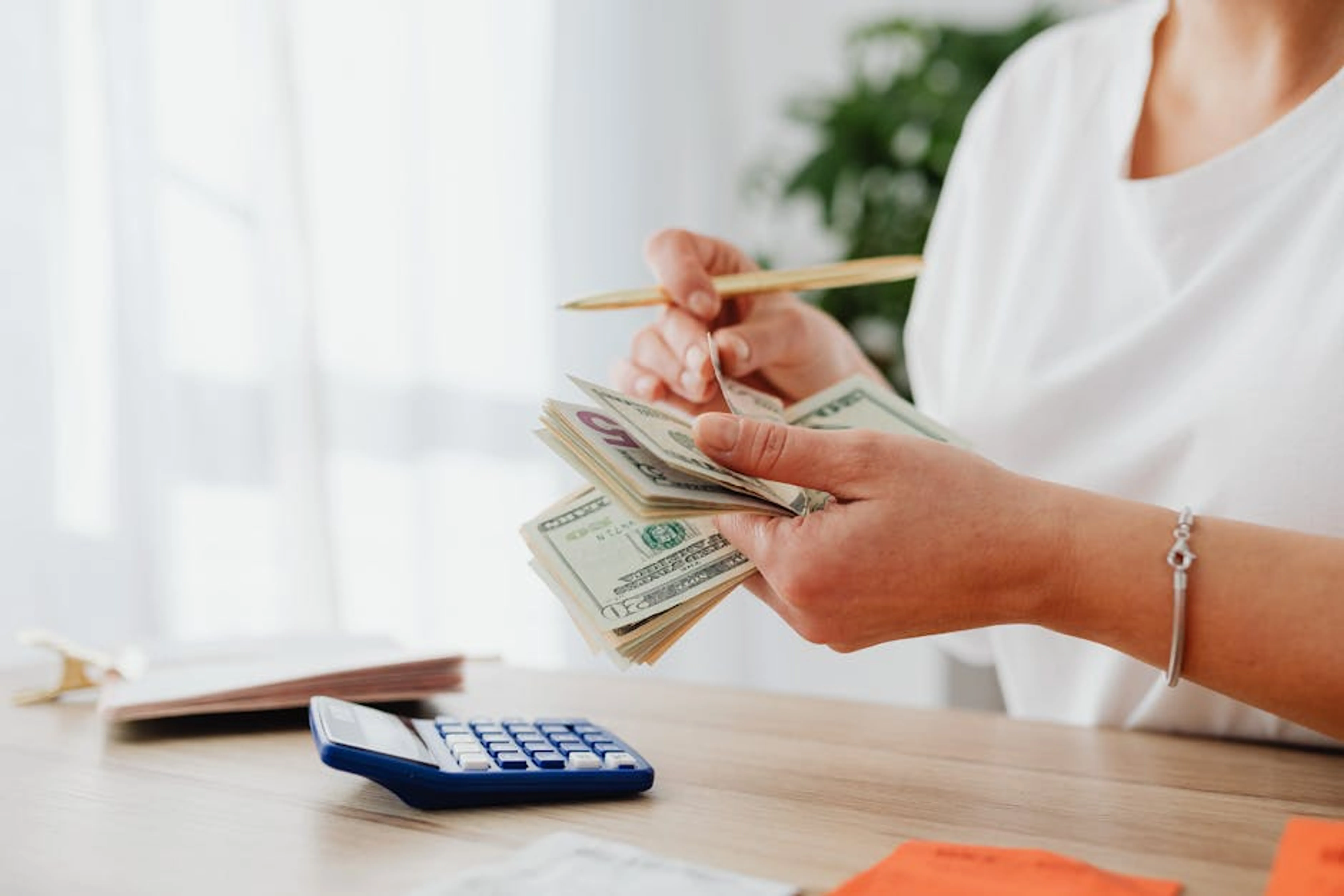 Close-up of a woman holding US dollars, calculating finances at home. A person reviewing financial documents on a tablet, with a calculator and coffee cup nearby
