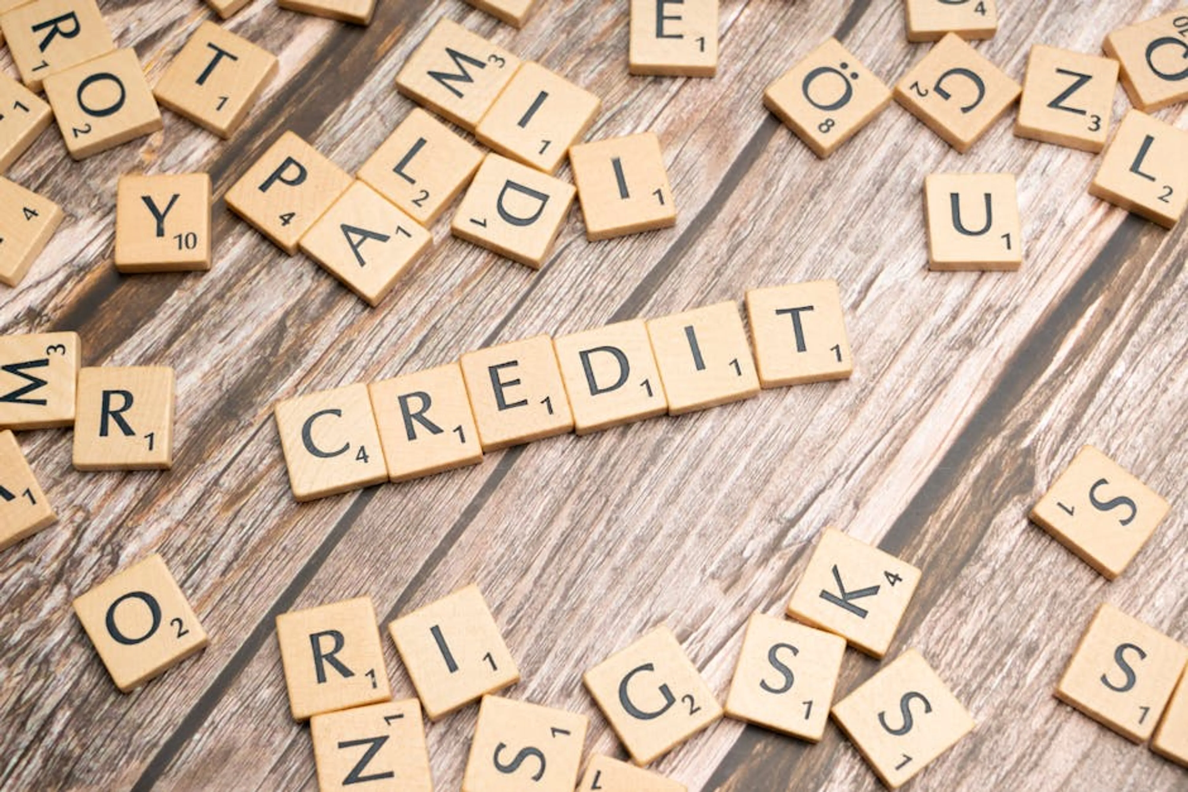 Wooden letter tiles arranged to spell 'CREDIT' on a rustic table background. Person holding a credit card and looking at a credit score chart