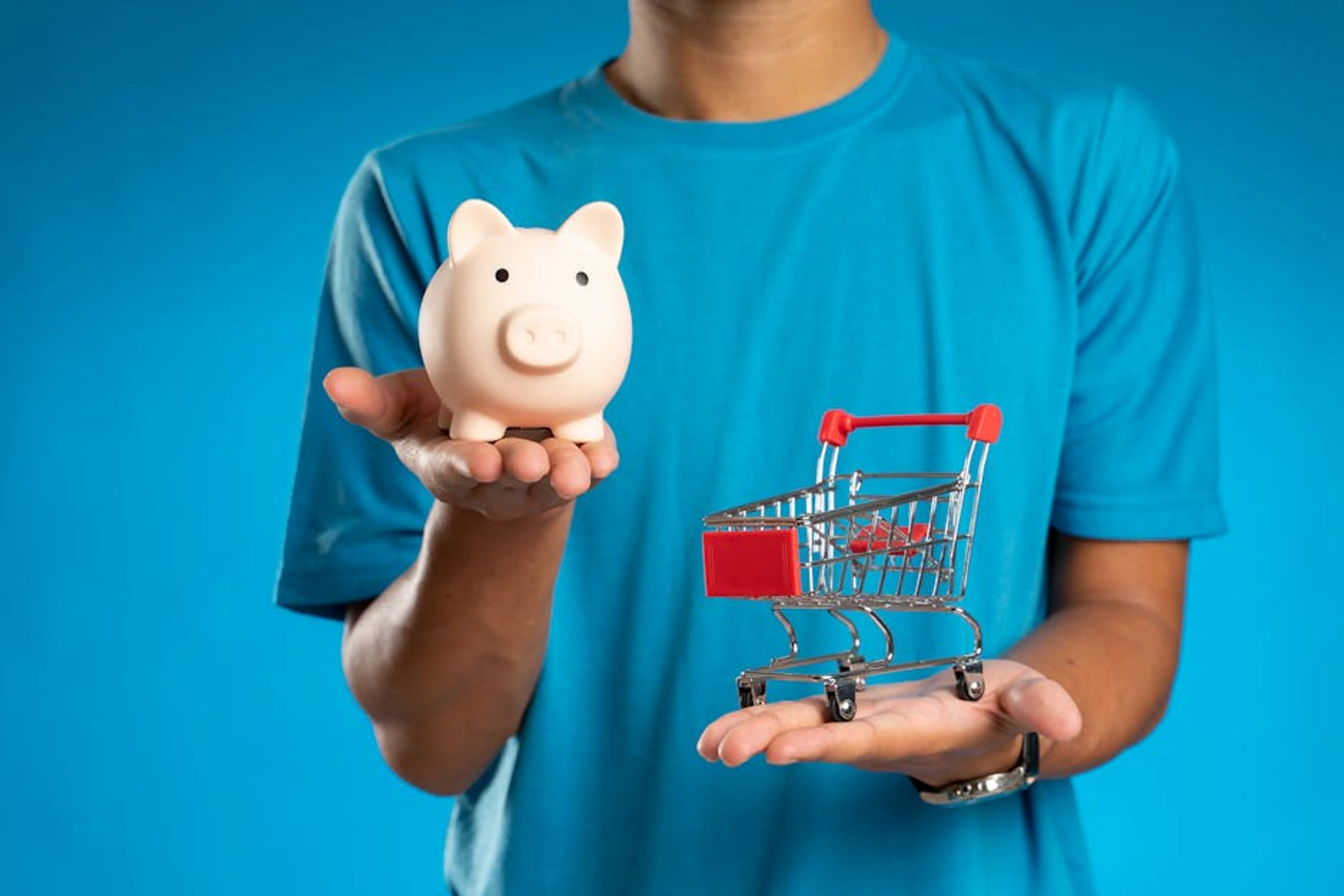 Man in blue shirt holding piggy bank and mini shopping cart, symbolizing savings versus spending. Person using a calculator and laptop to make financial decisions