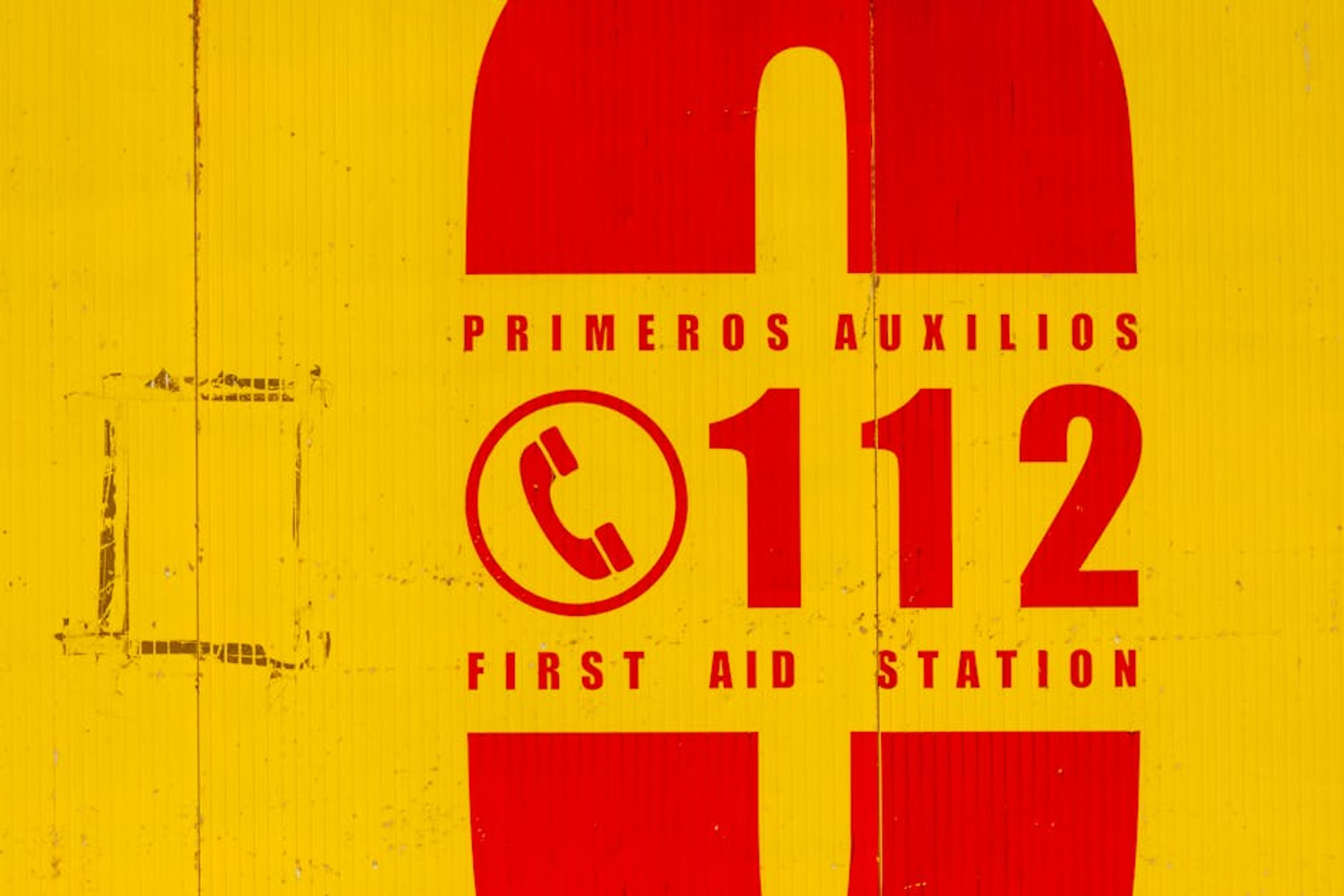 Bright yellow and red emergency sign indicating first aid station. Call 112 for emergencies. A hand holding a phone displaying emergency contact numbers and a map with embassy location.