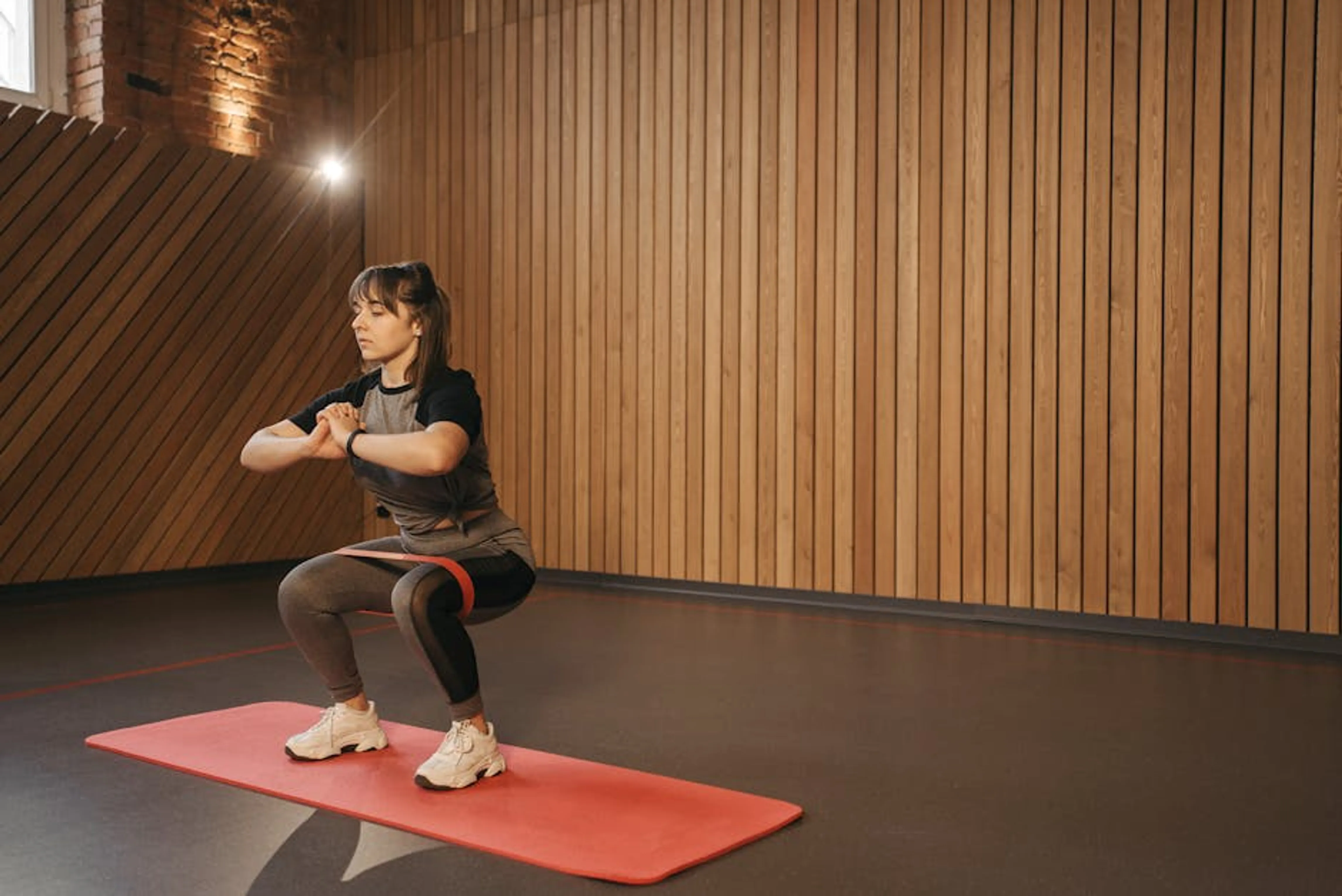Young woman exercises indoors with resistance band on mat for fitness Beginner performing a squat with good form in a gym mirror